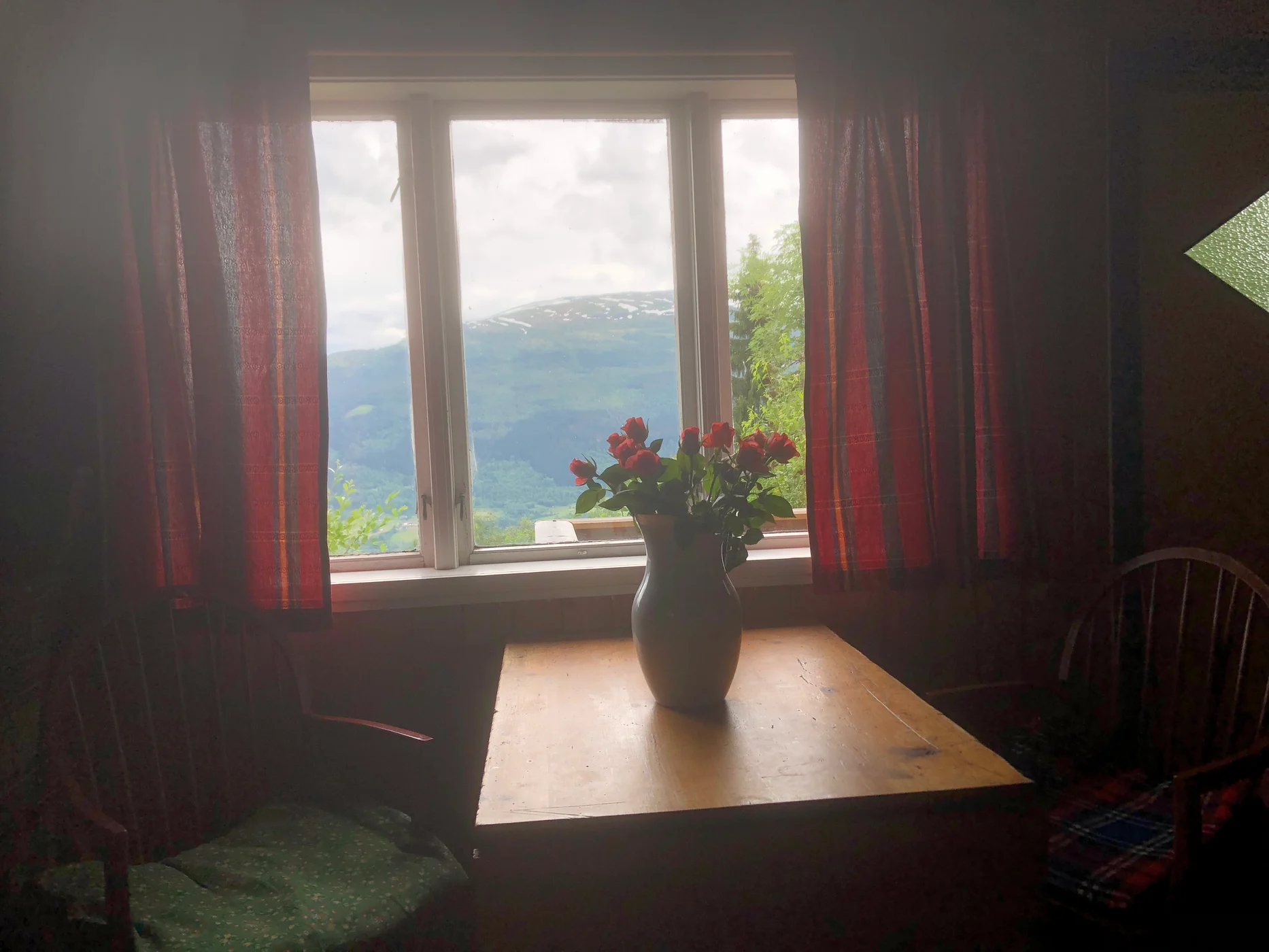 Vase med raude roser på trebord framfor vindauge med fjellutsikt
